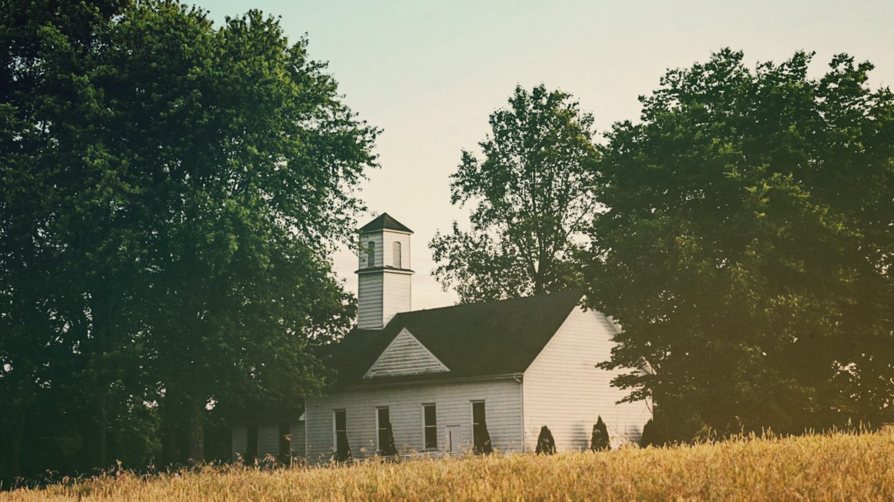 A small white country church with a square steeple, set among trees behind a field of ripe wheat in evening light.