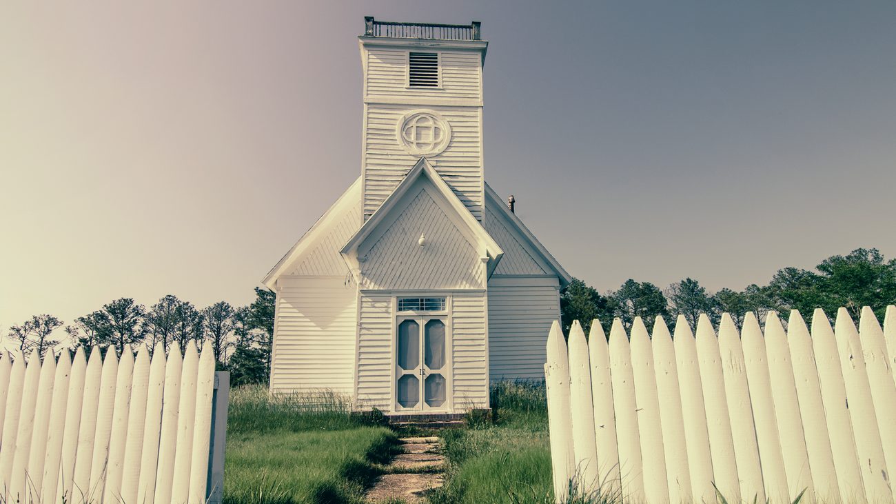 A small white clapboard country church with a square bell tower, seen through an open picket-fence gate with a worn footpath leading to its front door.