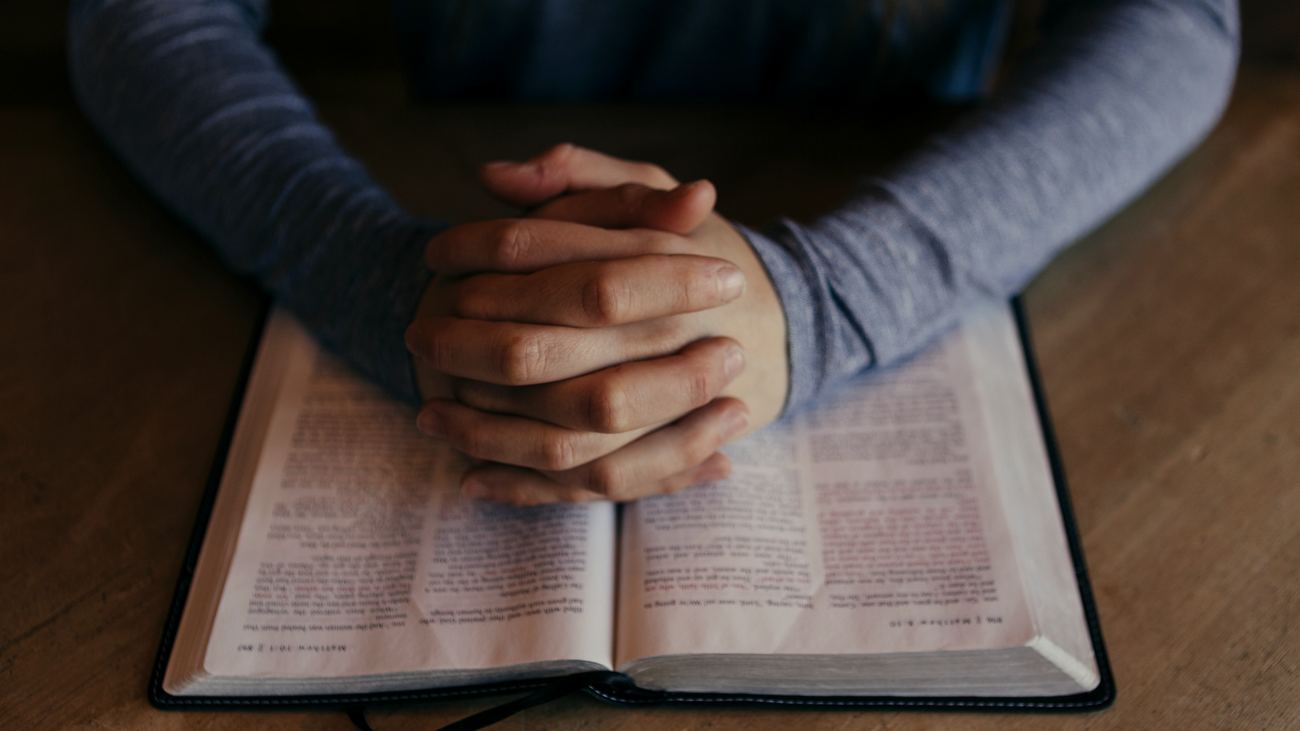 Hands folded on an open Bible on a wooden table.