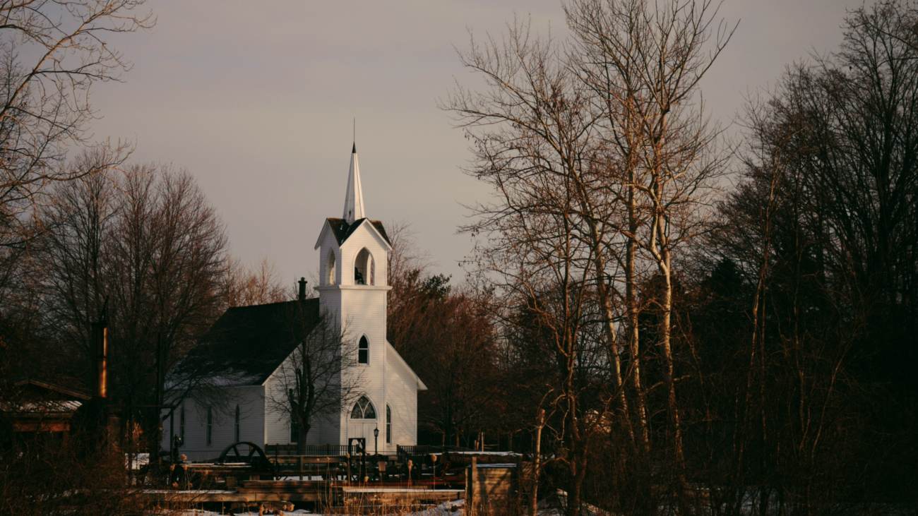 A small white-frame country church with a steeple, among bare trees in low golden evening light.
